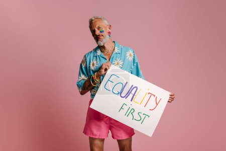 Téléchargez les photos : Confiant homme gay mature avec drapeau arc-en-ciel maquillage tenant bannière sur fond rose - en image libre de droit