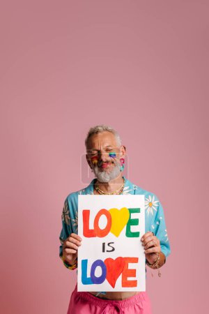 Foto de Feliz maduro gay hombre con arco iris bandera maquillaje mostrando colorido banner contra rosa fondo - Imagen libre de derechos