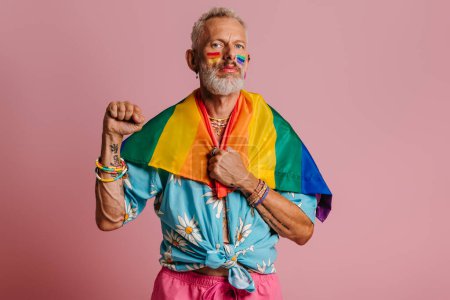 Téléchargez les photos : Confiant homme gay mature portant drapeau arc-en-ciel sur les épaules tout en se tenant sur fond rose - en image libre de droit