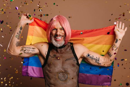 Foto de Alegre gay hombre en rosa peluca llevar arco iris bandera mientras confeti volando alrededor él contra marrón fondo - Imagen libre de derechos