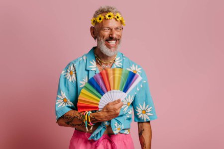 Photo for Fashionable mature gay man in floral wreath waving hand fan and smiling against pink background - Royalty Free Image
