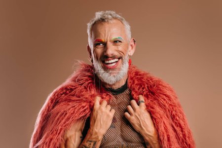 Téléchargez les photos : Heureux mature gay l'homme avec arc-en-ciel coloré sourcils portant peluche manteau sur brun fond - en image libre de droit