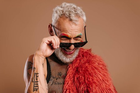 Téléchargez les photos : Élégant mature gay l'homme avec coloré sourcils ajustement lunettes et sourire sur brun fond - en image libre de droit
