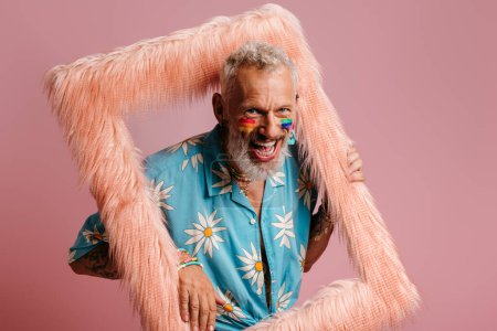 Foto de Alegre maduro gay hombre con arco iris bandera maquillaje buscando fuera de el esponjoso marco contra rosa fondo - Imagen libre de derechos