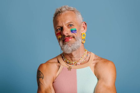 Téléchargez les photos : Portrait d'homme gay mature avec drapeau arc-en-ciel maquillage en regardant la caméra sur fond bleu - en image libre de droit