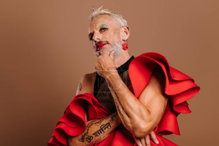 Foto de Confiado hombre gay maduro con corona en la cabeza usando hermoso vestido rojo contra fondo marrón - Imagen libre de derechos