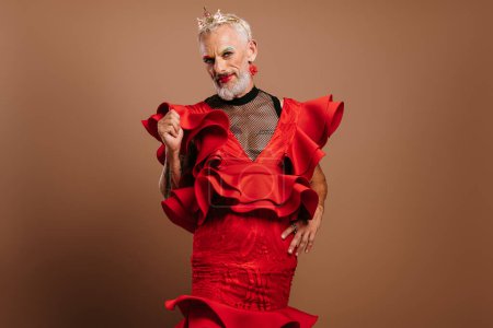 Foto de Barbudo maduro gay hombre con corona en cabeza usando hermosa rojo vestido contra marrón fondo - Imagen libre de derechos