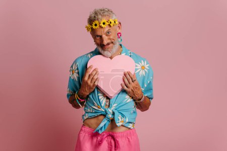Foto de Maduro gay hombre en floral guirnalda celebración corazón forma y sonriendo contra rosa fondo - Imagen libre de derechos