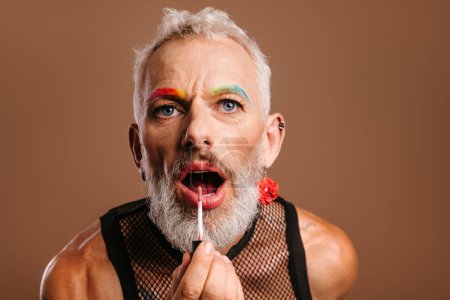 Foto de Barbudo maduro gay hombre con arco iris color cejas aplicando lápiz labial contra marrón fondo - Imagen libre de derechos