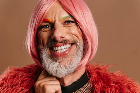 Foto de Retrato de hombre gay maduro con cejas de color arco iris usando peluca rosa contra fondo marrón - Imagen libre de derechos