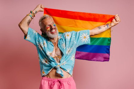 Téléchargez les photos : Joyeuse mature gay homme portant drapeau arc-en-ciel et souriant sur fond rose - en image libre de droit