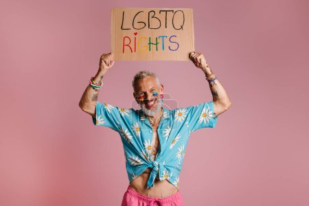 Photo for Happy mature gay man with rainbow flag make-up holding banner against pink background - Royalty Free Image