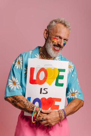 Foto de Alegre maduro gay hombre con arco iris bandera maquillaje celebración colorido banner contra rosa fondo - Imagen libre de derechos