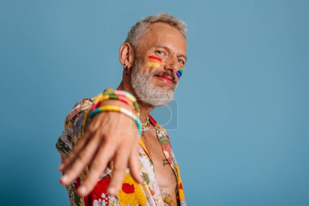 Téléchargez les photos : Mature gay homme avec arc-en-ciel drapeau maquillage étirant la main et souriant sur fond bleu - en image libre de droit