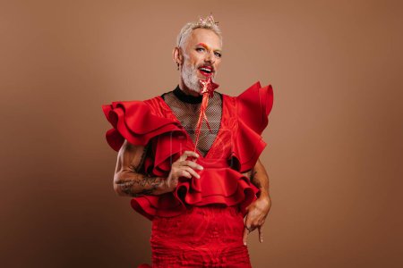 Téléchargez les photos : Homme gay ludique en belle robe rouge tenant baguette magique sur fond brun - en image libre de droit