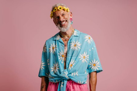 Téléchargez les photos : Heureux homme âgé en chemise élégante et couronne de fleurs sur la tête debout sur fond rose - en image libre de droit