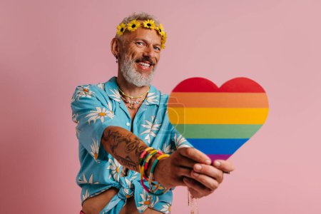 Photo for Happy mature gay man in floral wreath holding heart shaped rainbow flag against pink background - Royalty Free Image
