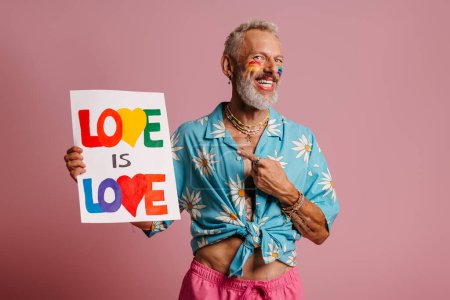 Foto de Feliz maduro gay hombre con arco iris bandera maquillaje apuntando colorido banner contra rosa fondo - Imagen libre de derechos