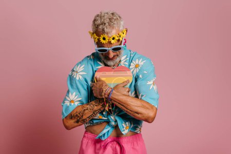 Téléchargez les photos : Mature gay l'homme dans floral couronne embrassant coeur en forme de drapeau arc-en-ciel sur rose fond - en image libre de droit
