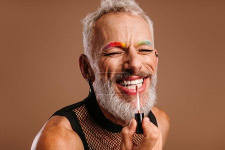 Foto de Feliz hombre gay maduro con sonrisa sincera aplicando maquillaje contra fondo marrón - Imagen libre de derechos