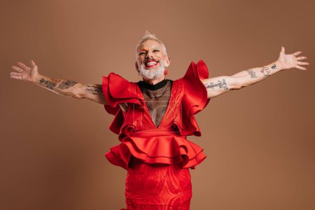 Photo for Joyful gay man with crown on head wearing beautiful red dress and gesturing against brown background - Royalty Free Image