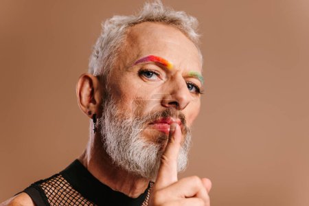 Foto de Barbudo maduro gay hombre con arco iris color cejas celebración dedo en labios contra marrón fondo - Imagen libre de derechos