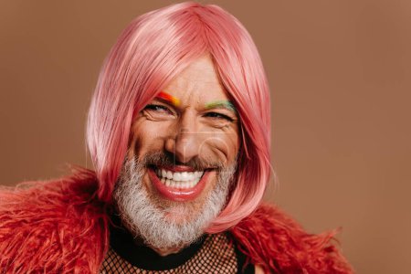 Foto de Feliz maduro gay hombre con arco iris color cejas usando rosa peluca contra marrón fondo - Imagen libre de derechos