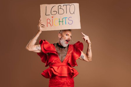 Téléchargez les photos : Mature gay homme dans belle robe rouge tenant bannière et pointant avec sourire sur fond brun - en image libre de droit