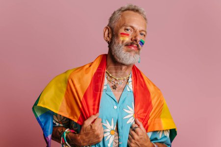 Photo for Confident mature gay man carrying rainbow flag on shoulders while standing against pink background - Royalty Free Image