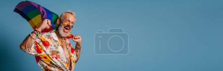 Photo for Happy mature gay man carrying rainbow flag and smiling while standing against blue background - Royalty Free Image