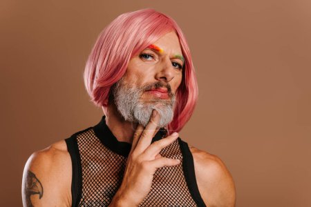 Foto de Retrato de hombre gay maduro con cejas de color arco iris usando peluca rosa contra fondo marrón - Imagen libre de derechos