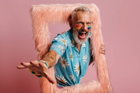 Téléchargez les photos : Ludique mature gay l'homme avec arc-en-ciel drapeau maquillage regarder hors de l 'moelleux cadre sur rose fond - en image libre de droit