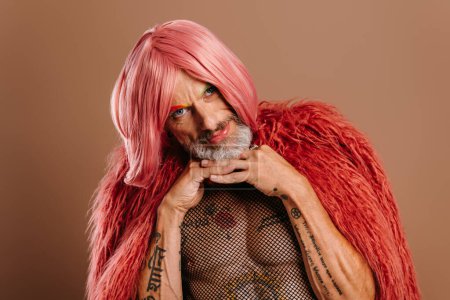 Téléchargez les photos : Barbu mature gay l'homme dans rose perruque penché visage sur les mains sur brun fond - en image libre de droit