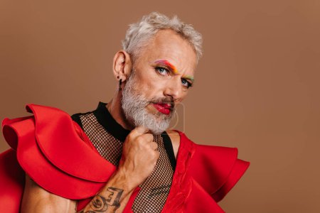 Téléchargez les photos : Portrait d'homme gay mature confiant en robe rouge regardant la caméra sur fond brun - en image libre de droit