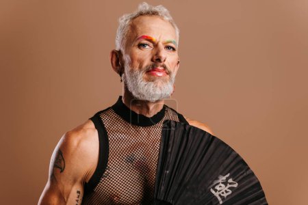 Foto de Barbudo maduro gay hombre con arco iris color cejas celebración mano ventilador contra marrón fondo - Imagen libre de derechos