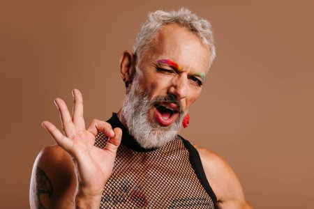 Foto de Juguetón maduro gay hombre con arco iris color cejas gesto y guiño contra marrón fondo - Imagen libre de derechos