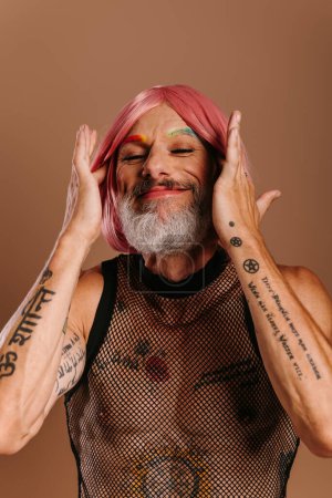 Foto de Feliz maduro gay hombre con arco iris color cejas ajuste su rosa peluca contra marrón fondo - Imagen libre de derechos