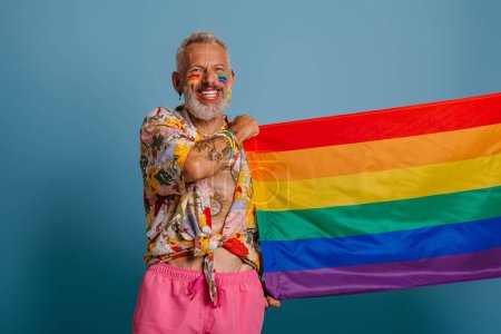 Foto de Alegre maduro gay hombre llevando arco iris bandera y sonriendo mientras de pie sobre azul fondo - Imagen libre de derechos