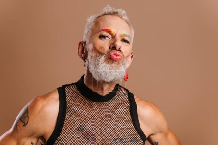 Téléchargez les photos : Portrait d'homme gay mature barbu avec maquillage soufflant un baiser tout en se tenant debout sur fond brun - en image libre de droit