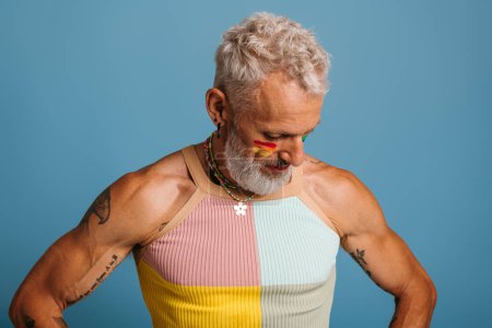 Téléchargez les photos : Mature gay l'homme avec arc-en-ciel drapeau maquillage regarder vers le bas alors que debout sur bleu fond - en image libre de droit