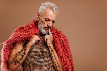 Téléchargez les photos : Portrait d'homme gay mature avec des sourcils colorés arc-en-ciel portant un manteau moelleux sur fond brun - en image libre de droit
