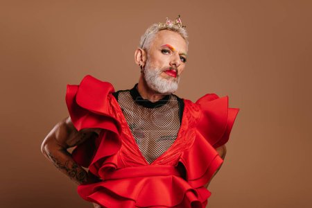 Téléchargez les photos : Portrait d'homme gay mature confiant en belle robe rouge regardant la caméra sur fond brun - en image libre de droit