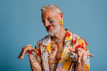 Photo for Mature gay man with rainbow flag make-up stretching out hand and smiling against blue background - Royalty Free Image