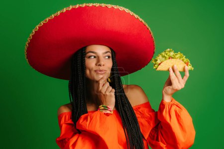 Foto de Atractiva joven mexicana en Sombrero sosteniendo taco y mirándolo con sonrisa sobre fondo verde - Imagen libre de derechos