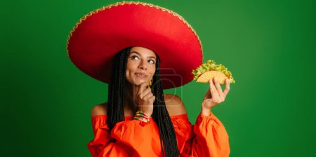 Photo for Beautiful young Mexican woman in Sombrero holding taco and looking thoughtful against green background - Royalty Free Image