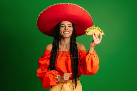 Photo for Attractive young Mexican woman in Sombrero holding taco and smiling against green background - Royalty Free Image