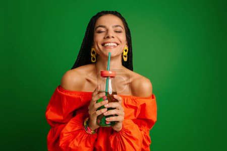 Téléchargez les photos : Joyeux jeune femme tenant bouteille avec de la limonade et gardant les yeux fermés sur fond vert - en image libre de droit