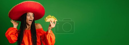 Photo for Attractive young Mexican woman in Sombrero holding taco and looking excited against green background - Royalty Free Image