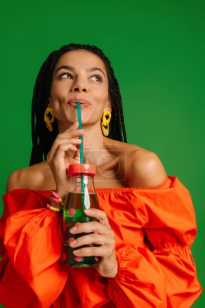 Téléchargez les photos : Jolie jeune femme appréciant la limonade fraîche et regardant heureux tout en se tenant debout sur fond vert - en image libre de droit