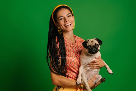 Foto de Joven mujer hispana alegre llevando un lindo perro pug y sonriendo sobre un fondo verde - Imagen libre de derechos